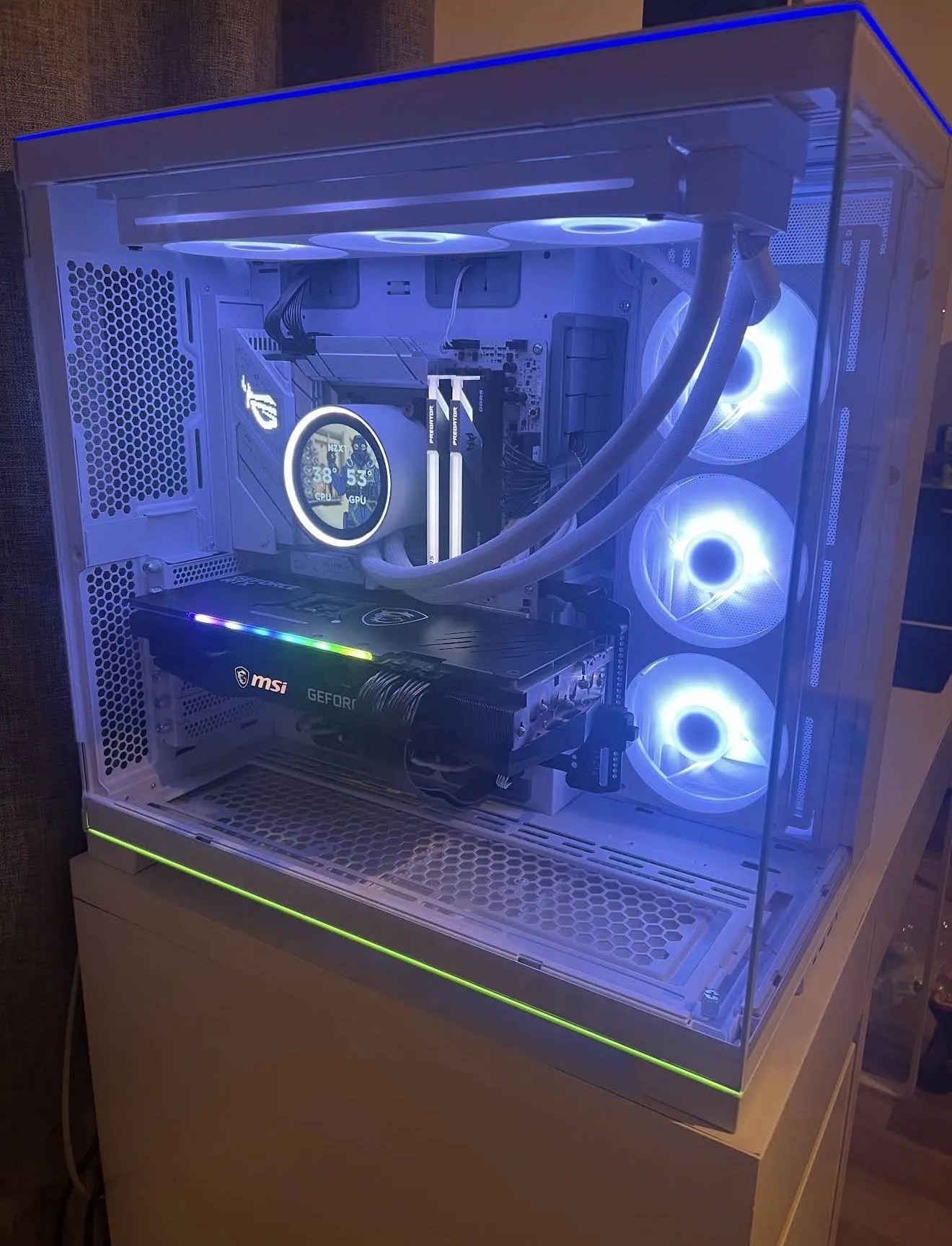 A white tempered-glass gaming PC lit in blue and white, with a liquid cooler showing live temperatures, RGB fans and a lit graphics card.