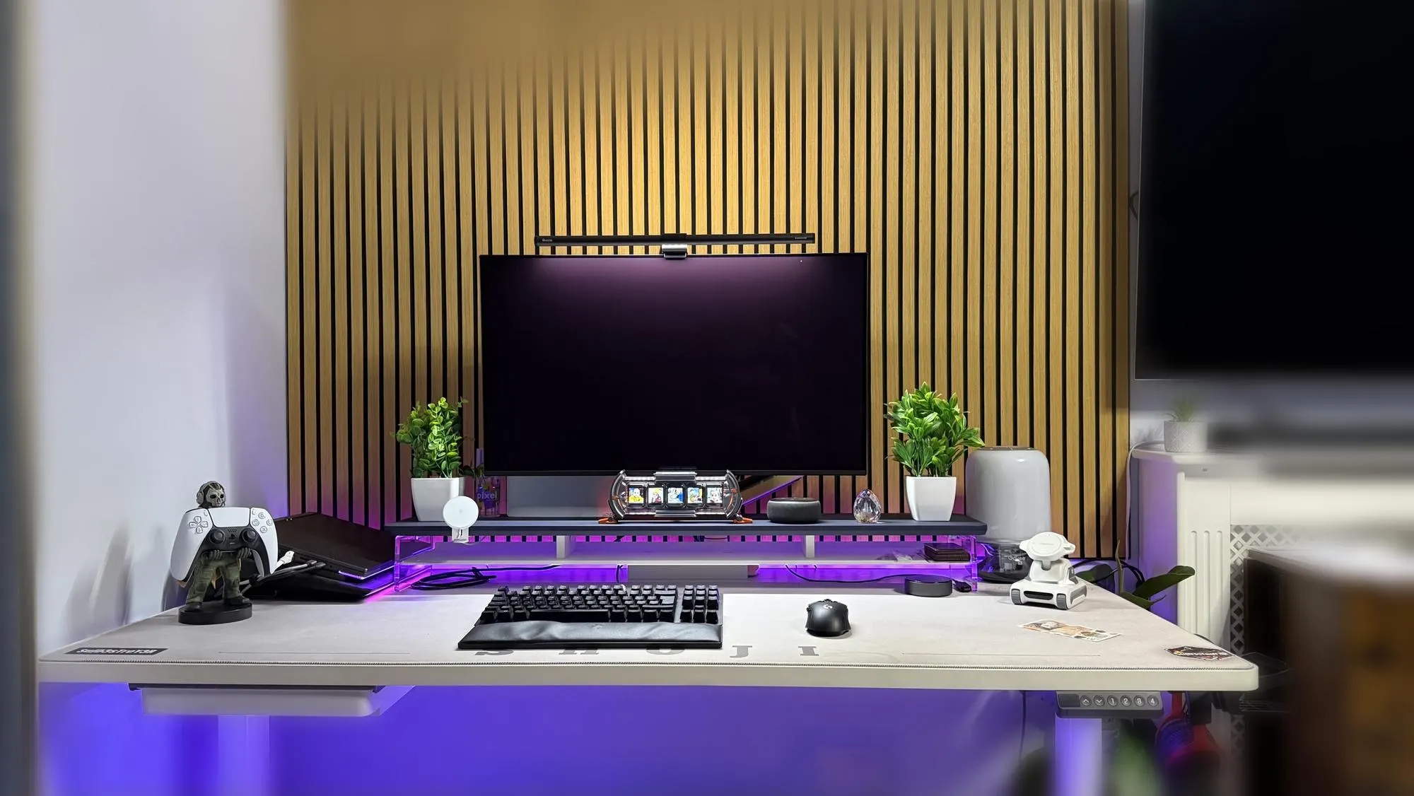 A tidy repair and planning desk with a large monitor, keyboard and mouse in front of a wooden slatted wall, lit with soft purple light